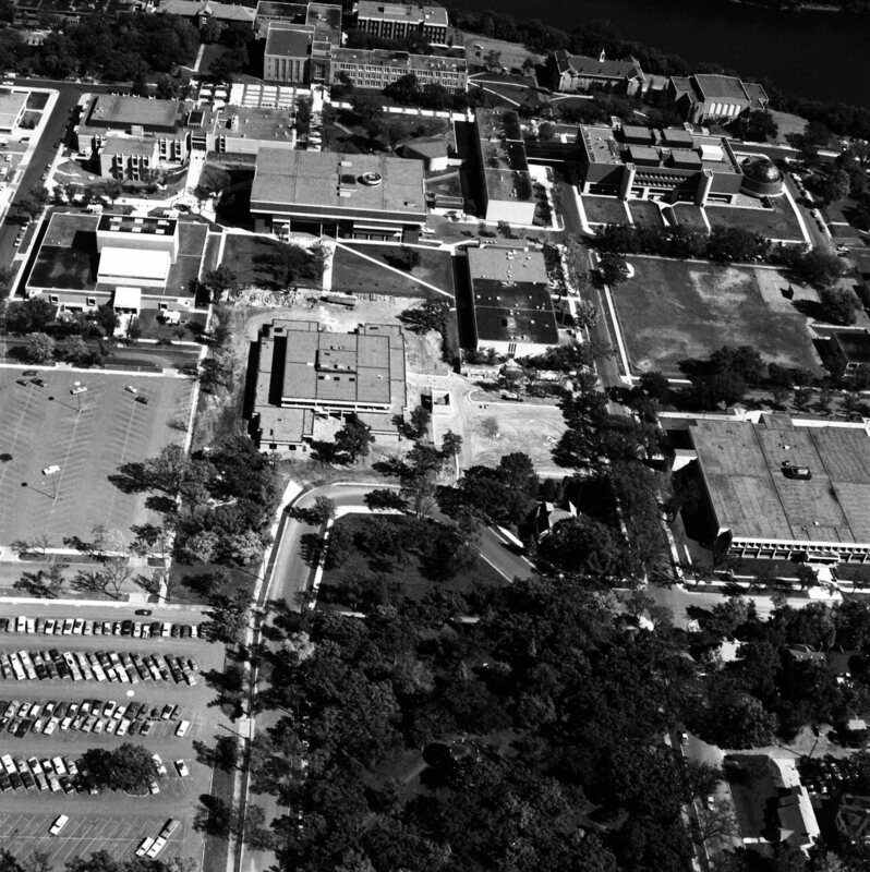Aerial black and white photograph of a university campus featuring multiple buildings of varying sizes and roof styles, surrounded by tree-lined pathways and parking lots filled with cars. A large open area with trees appears in the foreground, leading to a central building. A river runs horizontally across the background, framing the campus layout.
