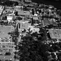 Aerial black and white photograph of a university campus featuring multiple buildings of varying sizes and roof styles, surrounded by tree-lined pathways and parking lots filled with cars. A large open area with trees appears in the foreground, leading to a central building. A river runs horizontally across the background, framing the campus layout.
